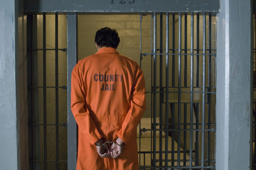 A man dressed in orange prison clothing stands before a jail cell